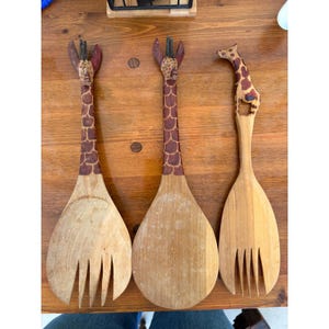 Puede incluir: Tres utensilios de ensalada de madera con mangos con temática de jirafa. Los mangos están pintados con patrones de jirafa marrones y beige. Los utensilios de ensalada están hechos de madera de color claro y tienen un diseño similar a un tenedor en el extremo.