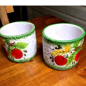 Puede incluir: Dos macetas de cerámica con base blanca y borde verde. Cada maceta presenta un diseño pintado a mano de manzanas rojas y hojas verdes. Las macetas son cilíndricas y están sobre una superficie de madera.