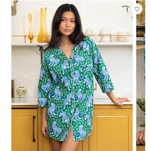 May include: A button-down sleep shirt featuring a vibrant green background with a repeating pattern of blue and white tigers. The garment has three-quarter length sleeves, a notched collar, and white piping trim along the front placket and cuffs.