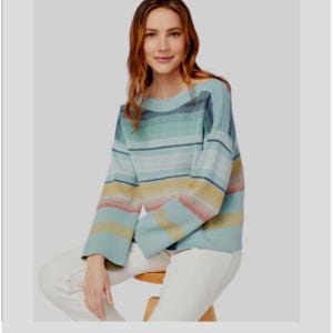 May include: A long-sleeve knit sweater featuring horizontal stripes in shades of teal, light blue, yellow, and soft pink. The sweater has a wide boat neckline and loose, relaxed sleeves, paired with plain white trousers.