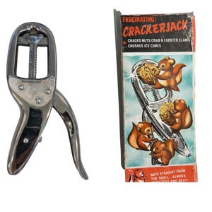 May include: A vintage Crackerjack nutcracker with a chrome finish, next to its original box. The box features illustrations of squirrels and text that reads "Crackerjack" and "Cracks Nuts-Crab & Lobster Claws Crushes Ice Cubes."