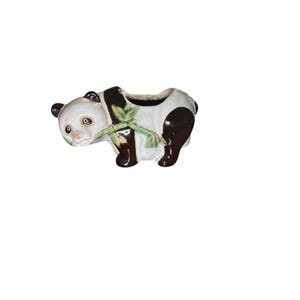 Puede incluir: Una maceta de cerámica con forma de oso panda. El panda tiene un cuerpo blanco, manchas negras y bambú verde. La maceta tiene una parte superior ahuecada para plantas. El panda sostiene una ramita de bambú.