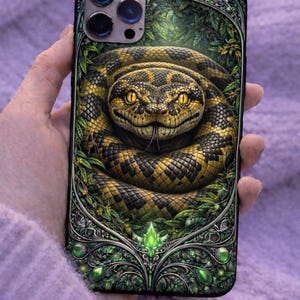 Puede incluir: Funda de teléfono con una ilustración detallada de una serpiente amarilla y negra enrollada en un exuberante entorno verde. La serpiente tiene ojos amarillos y una lengua bífida. La funda tiene un borde verde decorativo con acentos de gemas.