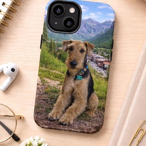 May include: A black and tan dog sits on a phone case. The case features a scenic mountain landscape with a town in the distance. The dog wears a blue collar with a tag. The case is on a light wood surface.