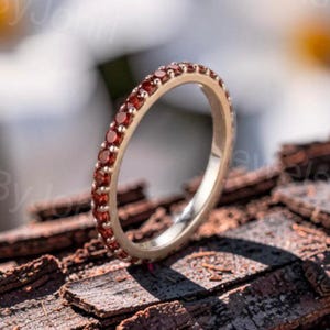 May include: A thin metal band ring featuring a continuous row of small, round, deep red garnet gemstones set along the outer circumference. The ring is positioned upright, showcasing the polished metal interior and the vibrant red stones.