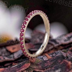 May include: A thin gold band ring featuring a continuous row of small, round, deep pink gemstones set along the outer circumference. The ring is positioned upright, showcasing the metallic finish and the uniform placement of the vibrant stones.