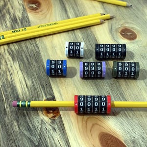 May include: A collection of six cylindrical mechanical row counters in white, black, blue, purple, and olive green, featuring rotating number wheels. One red counter is mounted on a yellow Ticonderoga pencil, with additional pencils nearby.