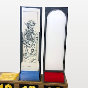 May include: Two vertical rectangular frames with dark gray borders. The left frame holds a white card featuring a black ink illustration of a goblin holding a dagger and a shield with a skull emblem, marked with a 0.75 inch measurement. The right frame is empty.