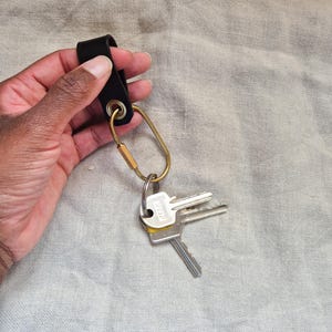 May include: A black leather key holder with a gold-colored carabiner clip and a set of keys. The key holder is attached to a gold-colored ring. The keys are silver and have yellow accents. The background is a light gray fabric.