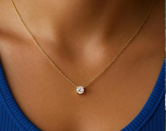 Può includere: Una delicata collana d'oro con un singolo pendente rotondo di pietra preziosa trasparente. Il pendente è centrato su una sottile catena d'oro. La collana è esposta su un top blu.