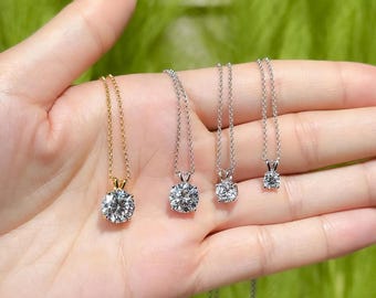 Puede incluir: Cuatro collares con colgantes de diamantes de diferentes tamaños. Un collar es dorado, los otros son plateados. Cada colgante presenta un diamante redondo y transparente. Los collares se muestran en una mano sobre un fondo verde borroso.