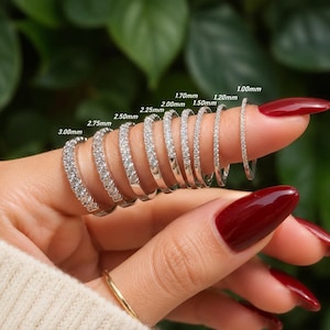 May include: A series of nine silver-toned eternity bands featuring clear stones, arranged by width from 1.00mm to 3.00mm. Each ring is labeled with its specific width measurement, showcasing a range of sizes from thin to thick bands.
