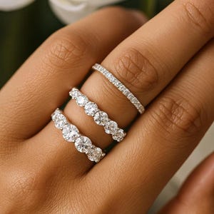 May include: Three diamond rings on a finger. The rings feature round-cut diamonds set in a silver band. One ring is a thin band of small diamonds, while the others have larger stones. The background is blurred.