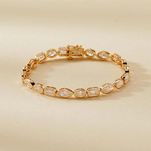 Könnte beinhalten: Ein goldfarbenes Armband mit klaren, rechteckigen und oval geformten Edelsteinen. Das Armband, mit einem sicheren Verschluss, wird vor einem neutralen Hintergrund präsentiert. Die Edelsteine sind gleichmäßig verteilt. Ein Schmuckstück.