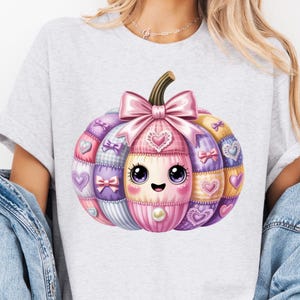 Peut inclure: T-shirt gris clair avec un motif de citrouille de dessin animé coloré. La citrouille est décorée de patchs roses, violets et jaunes, avec des cœurs et des nœuds. La citrouille a un visage mignon et un nœud rose sur le dessus.