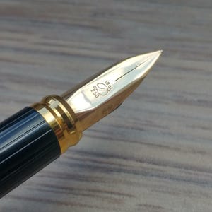 Puede incluir: Primer plano de una pluma estilogr&aacute;fica de lujo. La pluma tiene un plum&iacute;n dorado con detalles grabados y un cuerpo azul oscuro y dorado. El plum&iacute;n est&aacute; marcado con el n&uacute;mero 750 y otras marcas. Es un instrumento de escritura.