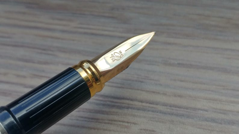 Puede incluir: Primer plano de una pluma estilogr&aacute;fica de lujo. La pluma tiene un plum&iacute;n dorado con detalles grabados y un cuerpo azul oscuro y dorado. El plum&iacute;n est&aacute; marcado con el n&uacute;mero 750 y otras marcas. Es un instrumento de escritura.