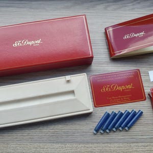 Puede incluir: Colecci&oacute;n de art&iacute;culos S.T. Dupont, que incluye una caja de cuero roja, un portal&aacute;pices blanco, un folleto, una tarjeta de garant&iacute;a, una peque&ntilde;a caja roja y cartuchos de tinta azules. Dispuestos sobre una superficie de madera.
