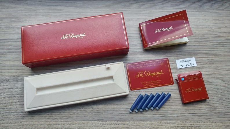 Puede incluir: Colecci&oacute;n de art&iacute;culos S.T. Dupont, que incluye una caja de cuero roja, un portal&aacute;pices blanco, un folleto, una tarjeta de garant&iacute;a, una peque&ntilde;a caja roja y cartuchos de tinta azules. Dispuestos sobre una superficie de madera.