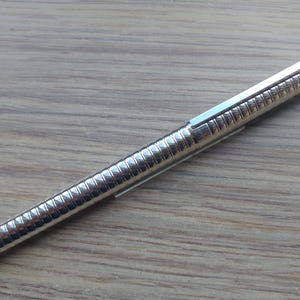 Puede incluir: Un bol&iacute;grafo plateado con un cuerpo acanalado y un clip rectangular. El bol&iacute;grafo est&aacute; en diagonal sobre una superficie de madera marr&oacute;n claro. El dise&ntilde;o del bol&iacute;grafo es elegante y moderno.