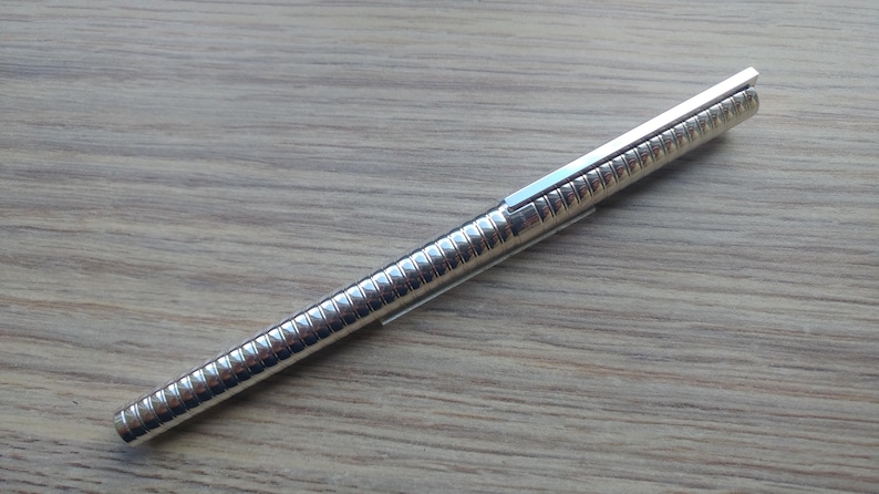 Puede incluir: Un bol&iacute;grafo plateado con un cuerpo acanalado y un clip rectangular. El bol&iacute;grafo est&aacute; en diagonal sobre una superficie de madera marr&oacute;n claro. El dise&ntilde;o del bol&iacute;grafo es elegante y moderno.