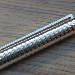 Puede incluir: Primer plano de un bol&iacute;grafo plateado brillante con un cuerpo texturizado y acanalado. El bol&iacute;grafo tiene un clip plateado en la parte superior. El bol&iacute;grafo est&aacute; sobre una superficie de madera.