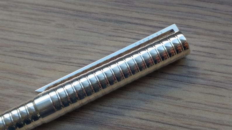Puede incluir: Primer plano de un bol&iacute;grafo plateado brillante con un cuerpo texturizado y acanalado. El bol&iacute;grafo tiene un clip plateado en la parte superior. El bol&iacute;grafo est&aacute; sobre una superficie de madera.