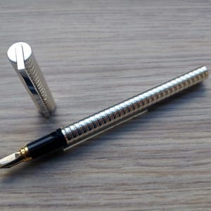 Puede incluir: Una pluma estilogr&aacute;fica plateada con un dise&ntilde;o acanalado, detalles en negro y un plum&iacute;n dorado. El capuch&oacute;n est&aacute; quitado y colocado junto a la pluma. La pluma descansa sobre una superficie de madera.