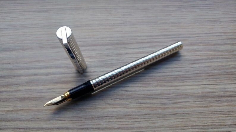 Puede incluir: Una pluma estilogr&aacute;fica plateada con un dise&ntilde;o acanalado, detalles en negro y un plum&iacute;n dorado. El capuch&oacute;n est&aacute; quitado y colocado junto a la pluma. La pluma descansa sobre una superficie de madera.