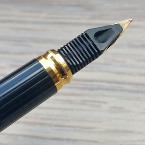 Puede incluir: Primer plano de una pluma estilogr&aacute;fica negra con detalles dorados. La pluma tiene un plum&iacute;n dorado y una secci&oacute;n de agarre negra y acanalada. El cuerpo de la pluma es negro con una banda dorada cerca del plum&iacute;n.