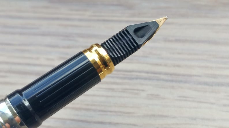 Puede incluir: Primer plano de una pluma estilogr&aacute;fica negra con detalles dorados. La pluma tiene un plum&iacute;n dorado y una secci&oacute;n de agarre negra y acanalada. El cuerpo de la pluma es negro con una banda dorada cerca del plum&iacute;n.