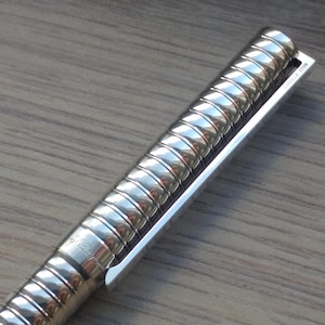 Puede incluir: Primer plano de un bol&iacute;grafo plateado con un cuerpo texturizado y acanalado y un clip plateado. El bol&iacute;grafo est&aacute; en &aacute;ngulo, mostrando los detalles del dise&ntilde;o. El bol&iacute;grafo est&aacute; sobre una superficie de madera.
