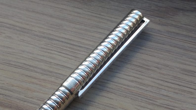 Puede incluir: Primer plano de un bol&iacute;grafo plateado con un cuerpo texturizado y acanalado y un clip plateado. El bol&iacute;grafo est&aacute; en &aacute;ngulo, mostrando los detalles del dise&ntilde;o. El bol&iacute;grafo est&aacute; sobre una superficie de madera.