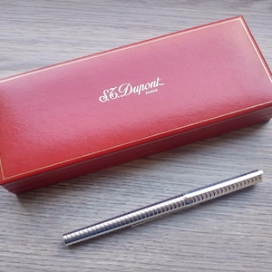 Puede incluir: Una caja rectangular roja con ribete dorado y el nombre de la marca "S.T. Dupont" en letras blancas. Un bol&iacute;grafo plateado con un dise&ntilde;o acanalado est&aacute; junto a la caja. El bol&iacute;grafo y la caja est&aacute;n sobre una superficie de madera.