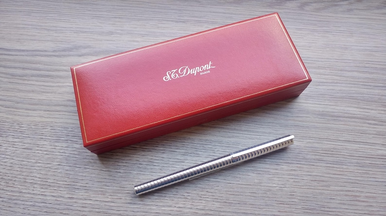 Puede incluir: Una caja rectangular roja con ribete dorado y el nombre de la marca "S.T. Dupont" en letras blancas. Un bol&iacute;grafo plateado con un dise&ntilde;o acanalado est&aacute; junto a la caja. El bol&iacute;grafo y la caja est&aacute;n sobre una superficie de madera.