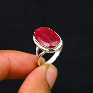 Może przedstawiać: Srebrny pierścionek z dużym, owalnym rubinem. Rubin ma głęboki, czerwony kolor i jest osadzony w polerowanej srebrnej oprawie. Projekt pierścionka jest prosty, ale elegancki, skupiając się na pięknie kamienia szlachetnego.