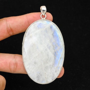 Puede incluir: Colgante de piedra lunar ovalado con una fianza de plata. La piedra lunar tiene una base blanca lechosa con destellos iridiscentes azules y morados. El colgante está engastado en un bisel de plata.