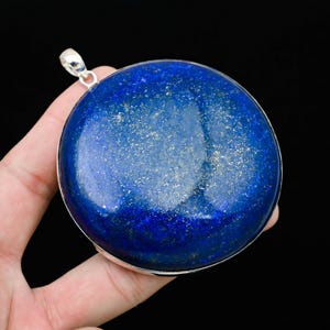 May include: A large, round, deep blue pendant with silver-colored trim and a loop for a chain. The pendant has a polished surface with flecks of gold, set against a black background. The pendant is held in a hand.