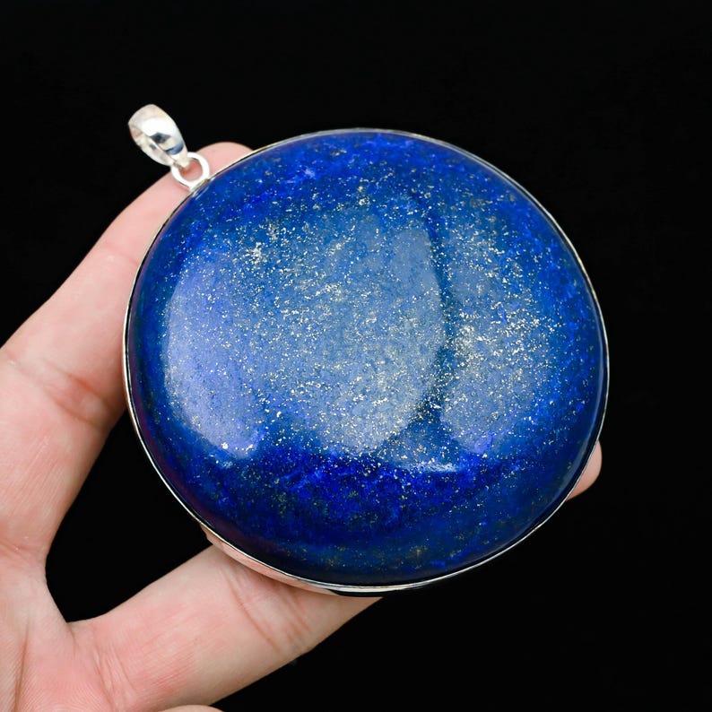 May include: A large, round, deep blue pendant with silver-colored trim and a loop for a chain. The pendant has a polished surface with flecks of gold, set against a black background. The pendant is held in a hand.