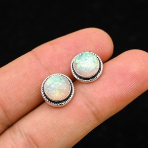 Peut inclure: Une paire de boucles d'oreilles à clous rondes en opale. Chaque boucle d'oreille présente une pierre d'opale irisée sertie dans une lunette argentée perlée. Les opales affichent des teintes blanches, vertes et jaunes, créant un jeu de couleurs subtil.