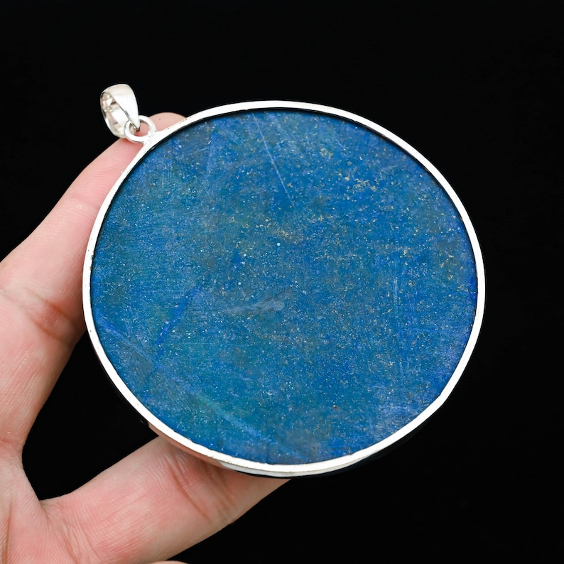 May include: A large, round pendant with a vibrant blue stone set in a silver-colored metal frame. The stone has natural variations in color and texture. The pendant has a small loop at the top for a chain.