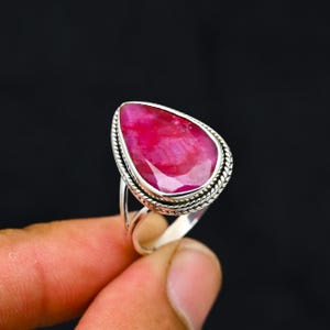 May include: A silver ring featuring a large, pear-shaped ruby gemstone. The ruby is a vibrant shade of red with visible internal patterns. The ring's setting has a textured, rope-like border around the gemstone.