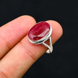 Könnte beinhalten: Ein Silberring mit einem großen, oval geschliffenen Rubin. Der Rubin hat eine tiefe rote Farbe und ist in einem schlichten, eleganten Silberband gefasst. Der Ring wird vor schwarzem Hintergrund gezeigt.