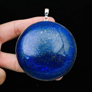 May include: A large, round, deep blue pendant with silver-colored trim and a silver bail. The pendant has a polished, reflective surface with flecks of gold. The gemstone is held in a hand against a black background.