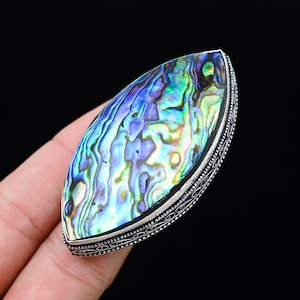 May include: A large, oval-shaped ring featuring a vibrant abalone shell with iridescent blues, greens, and purples. The shell is set in a silver-toned metal with a detailed, patterned border. The ring is held by a hand against a black background.