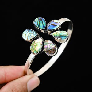 Pode incluir: Uma pulseira de punho prateada com seis pedras de concha abalone em forma de lágrima dispostas num design floral. As pedras iridescentes exibem tons de azul, verde e roxo. A pulseira está sobre um fundo preto.