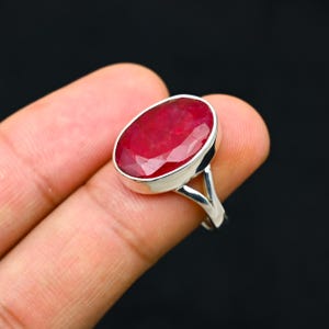 Puede incluir: Anillo de plata con una gran gema de rubí de corte ovalado. El rubí es de color rojo intenso y está engastado en una banda de plata pulida. El diseño del anillo es sencillo, centrado en la belleza de la gema.
