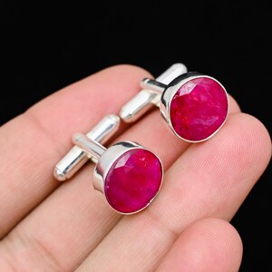May include: A pair of silver cufflinks with large, round, faceted ruby gemstones. The rubies are a vibrant, deep red color, set in a polished silver bezel. The cufflinks are displayed against a neutral background.