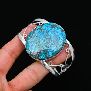 May include: A silver cuff bracelet featuring a large, oval turquoise stone with brown matrix patterns. The stone is set in a polished silver frame, and the bracelet has an open design with decorative cutouts. The background is black.