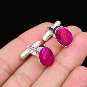 May include: A pair of silver cufflinks featuring oval, bright pink gemstones. The gemstones have a textured appearance and are set in a polished silver frame. The cufflinks are displayed against a dark background.